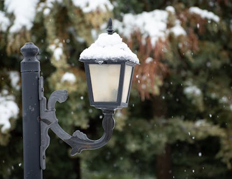 A snow-covered vintage street lamp illuminates a serene winter park scene with falling snowflakes.