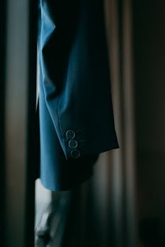Close-up of a stylish navy blue suit jacket hanging near a window in a softly lit room.