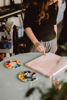 A woman artist paints on a canvas with a brush in a sunlit home studio, surrounded by vibrant paint palettes.