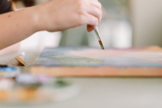 A female artist's hand skillfully painting on a canvas in a sunlit home studio setting.