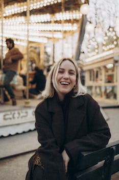 Blonde woman smiles happily in the foreground of a Parisian illuminated carousel at night.
