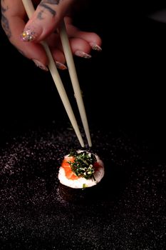 Close-up artistic photograph of sushi being held by chopsticks, adorned hand visible.