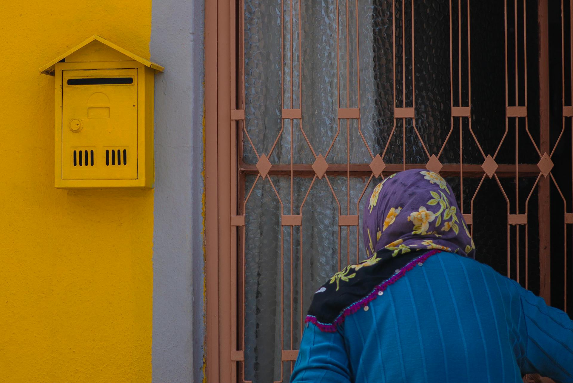 A woman in a floral scarf near a yellow mailbox and metal gate. Bright colors.