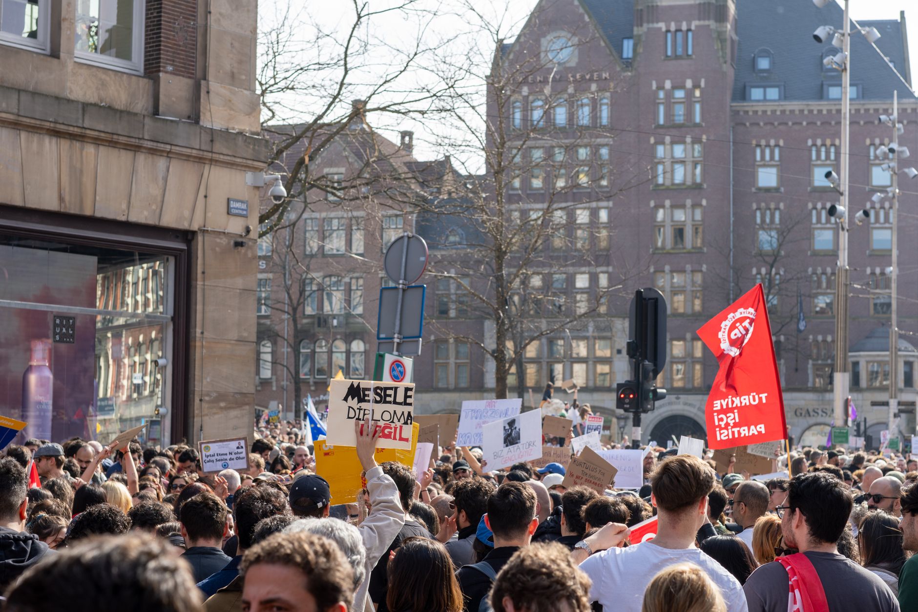 Een levendig stadsprotest met een diverse menigte die verschillende borden en spandoeken vasthoudt op een zonnige dag.