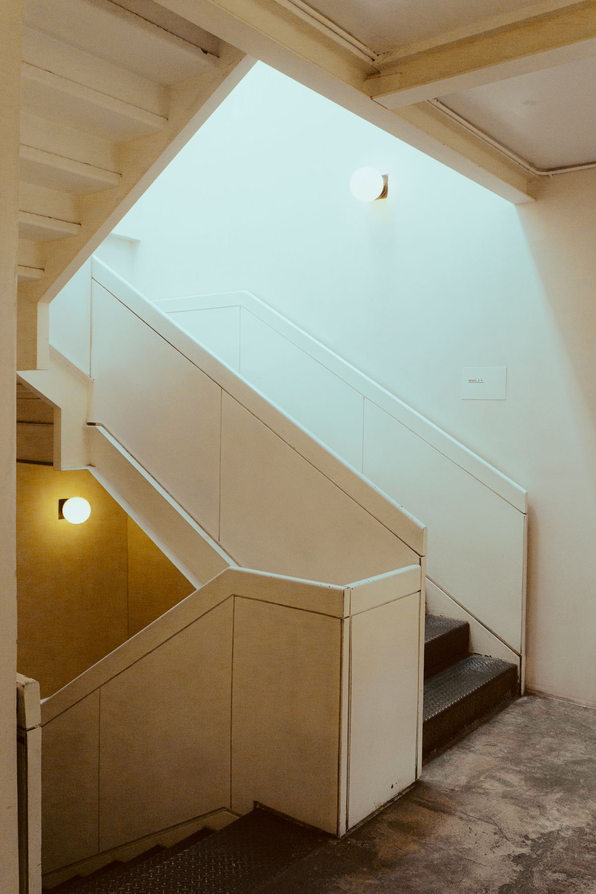Stairwell with pale plaster walls, warm wall lights and daylight falling from a skylight