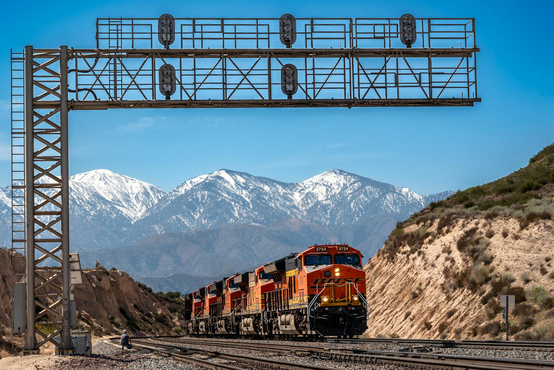 A vibrant freight train travels through a mountain range under blue skies, featuring snow-capped peaks.