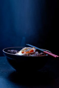 A flavorful Asian noodle bowl topped with beef, egg, and vegetables, artfully presented with chopsticks.