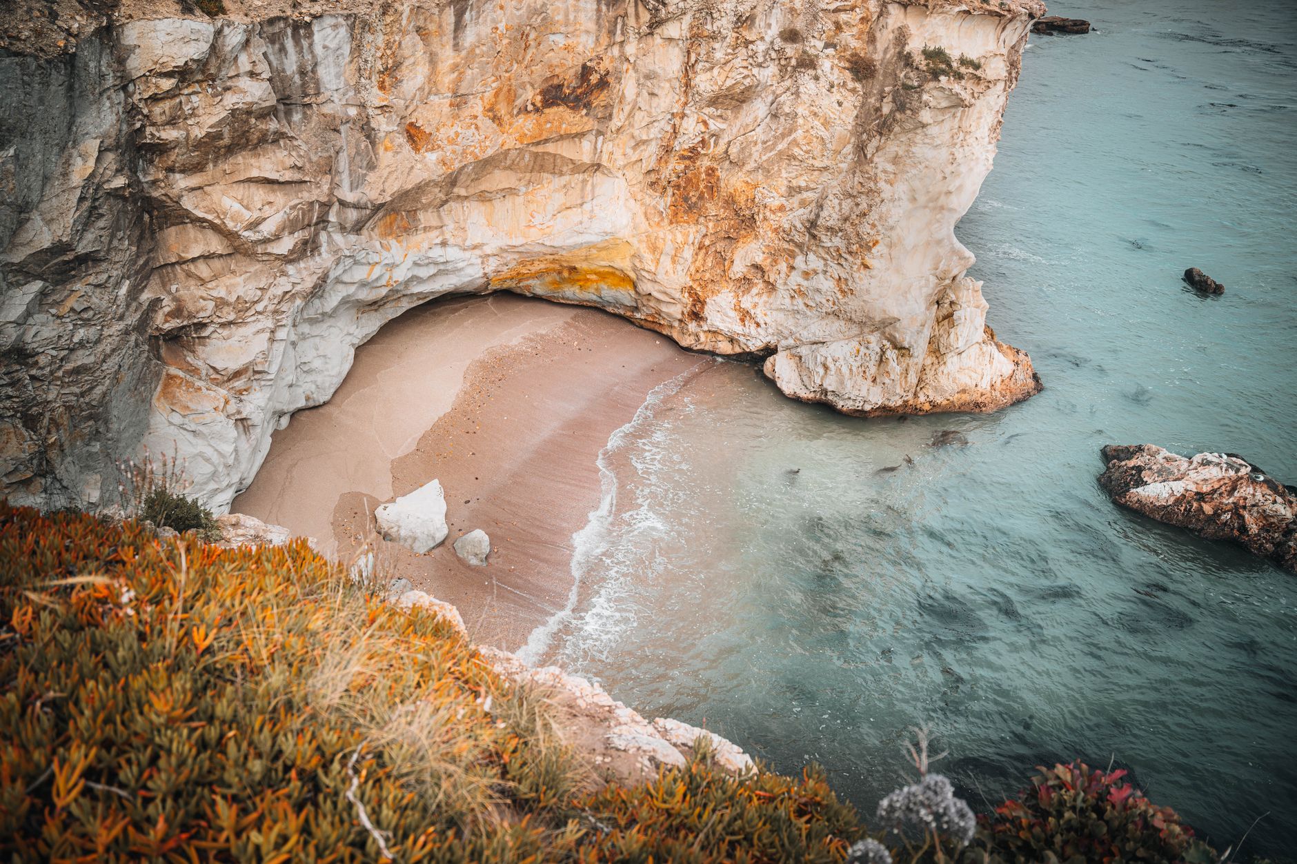 Aerial view of a serene cove with a sandy beach nestled beneath rugged cliffs, surrounded by azure water.