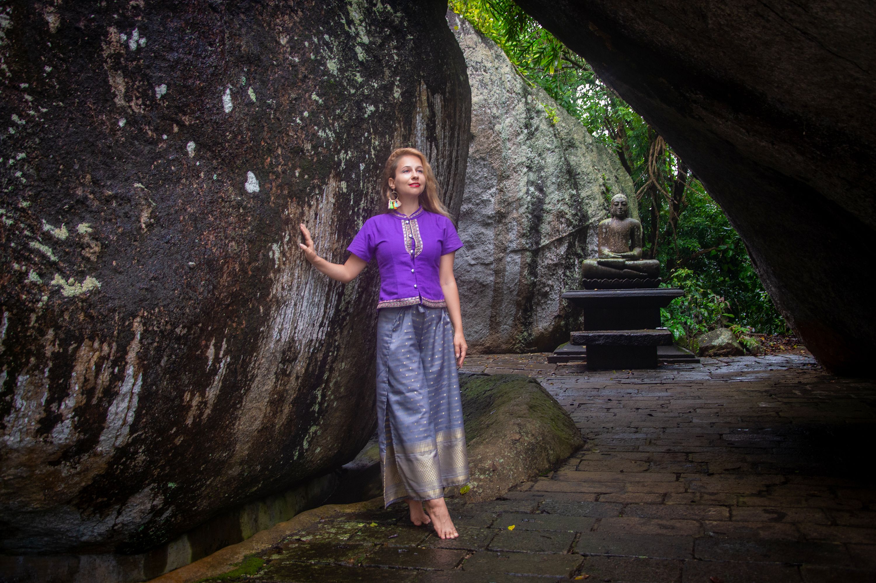 A woman in traditional clothing stands in a serene rock temple with a Buddha statue nearby.