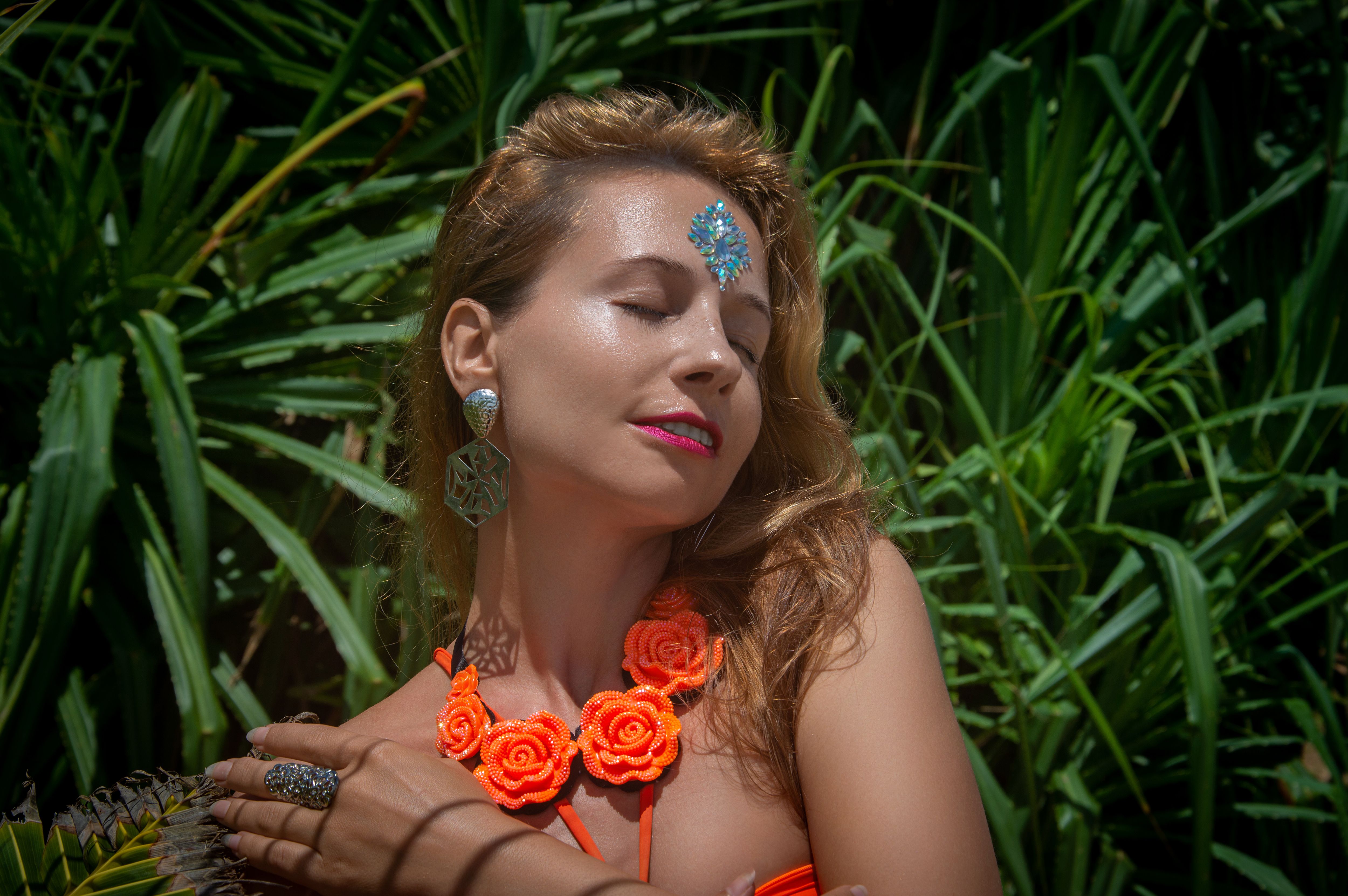 Woman enjoying sunlight with decorative jewelry, surrounded by lush green plants, exuding tranquility.
