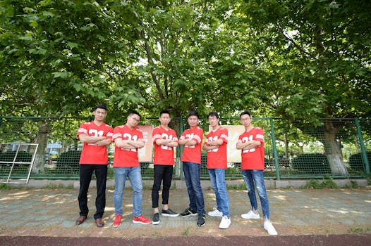Six young men in matching red jerseys pose confidently outdoors under green trees.
