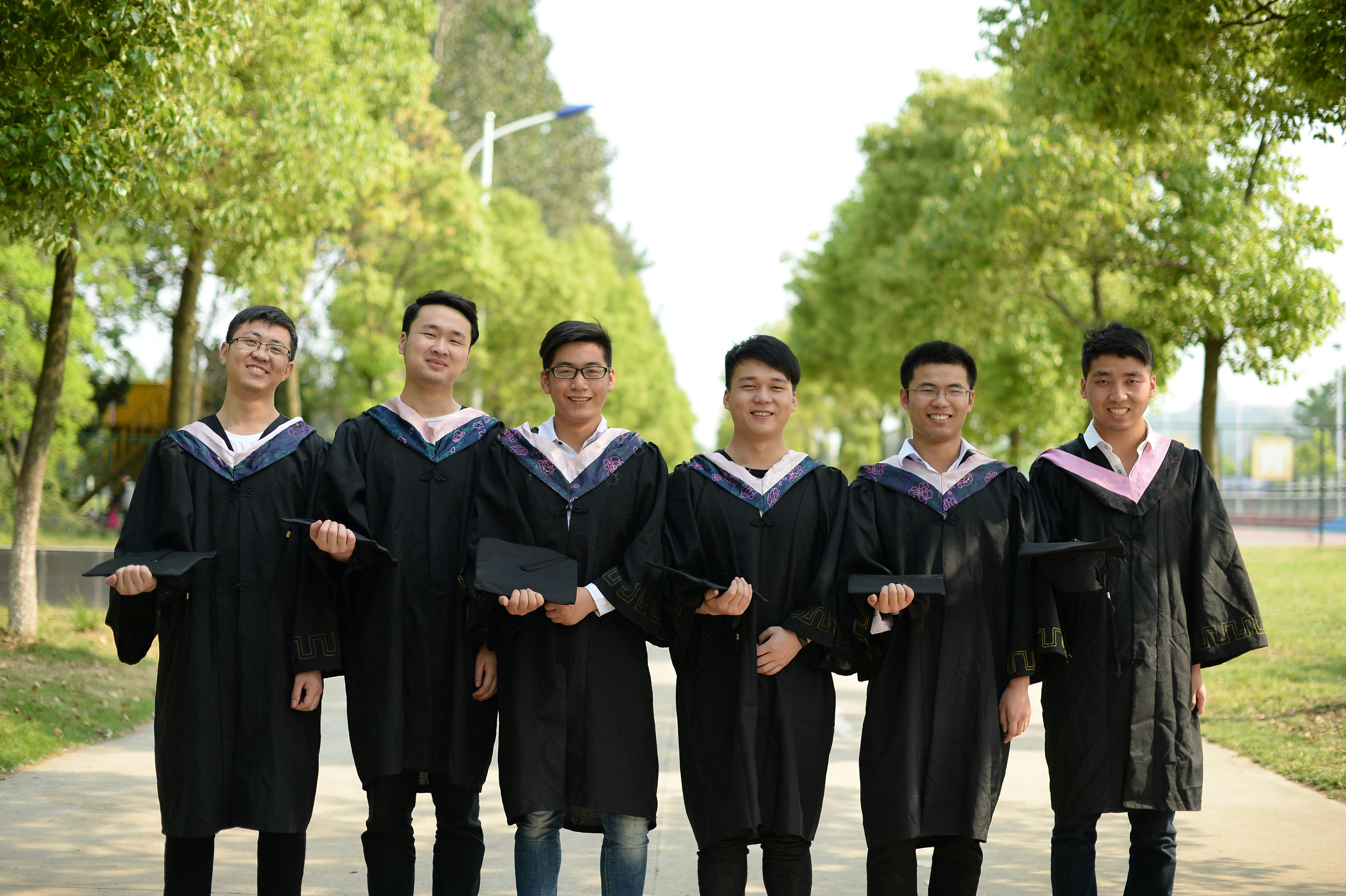 Six young graduates in gowns smiling and holding caps outdoors on a sunny day.