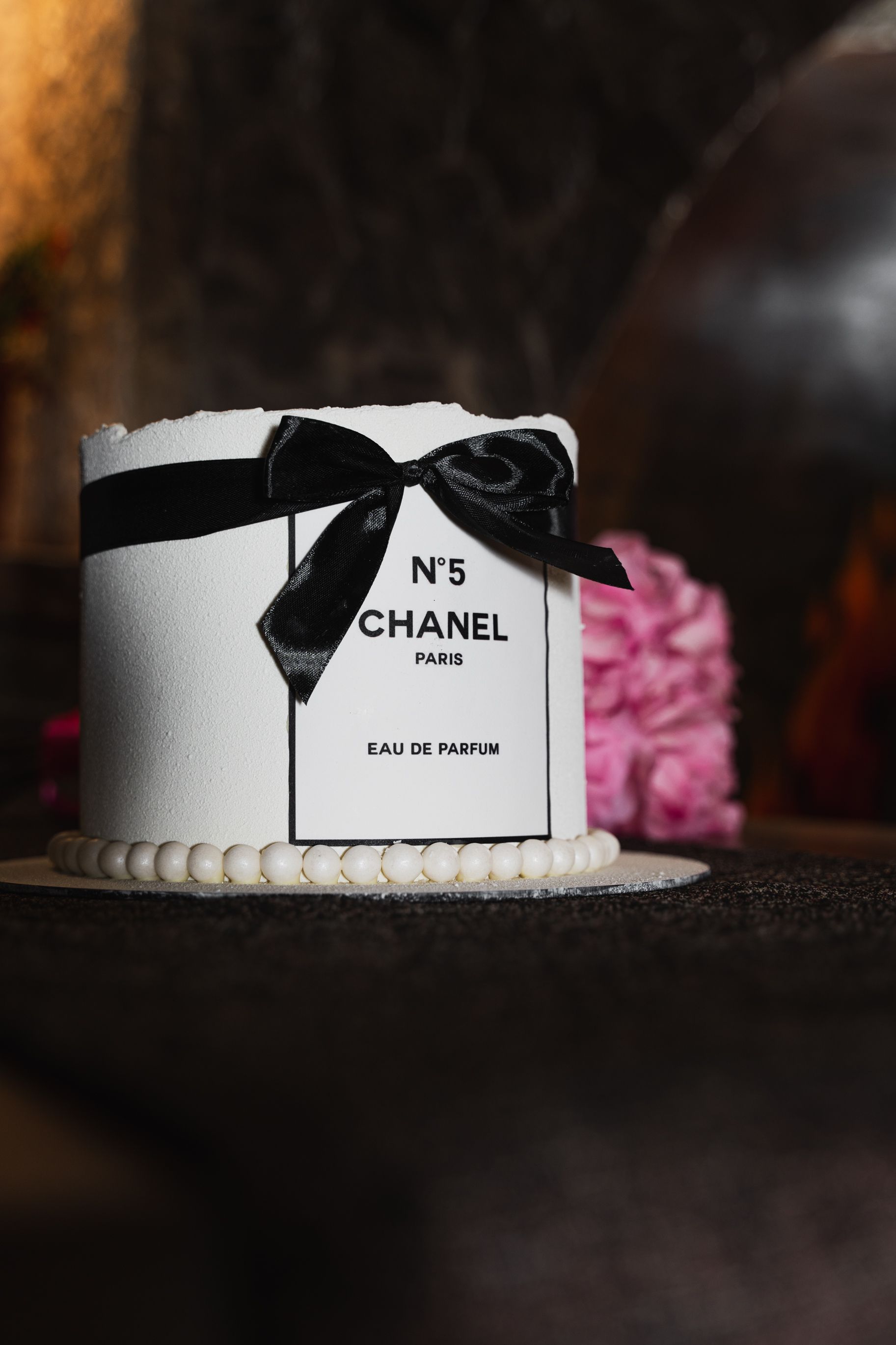 Um bolo temático Chanel No. 5, lindamente elaborado com um elegante laço preto em exibição, perfeito para eventos de luxo.