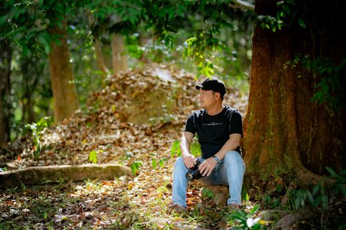 Free A man relaxes under a large tree in a serene forest, holding a camera and enjoying nature. Stock Photo