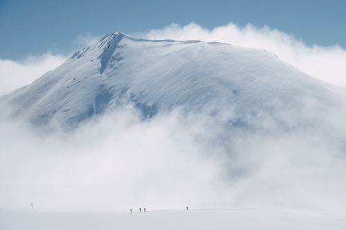 Free Snow-covered mountain peak enveloped by misty clouds with hikers below, showcasing winter adventure. Stock Photo