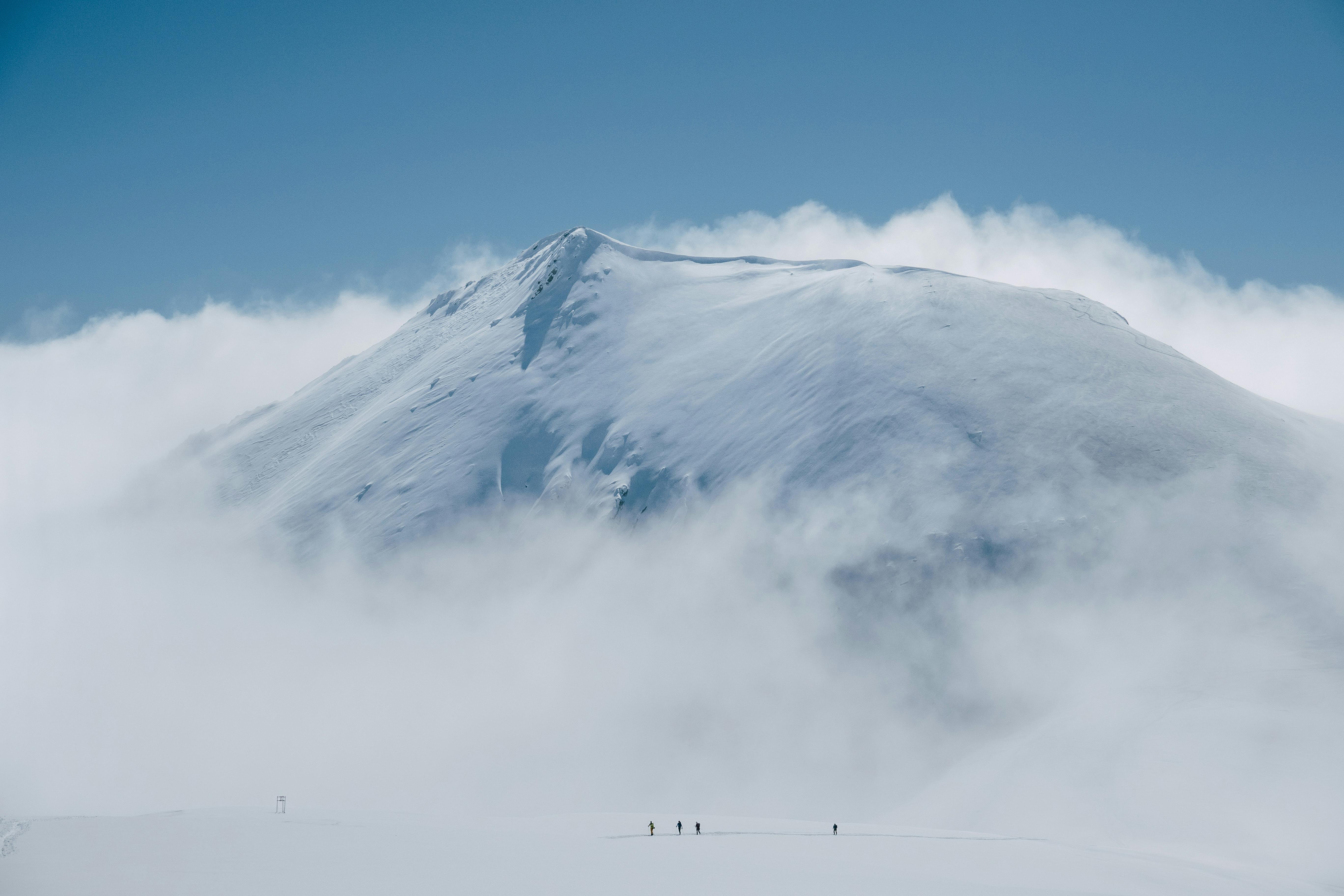 A breathtaking view of a snowy mountain surrounded by clouds with distant hikers.