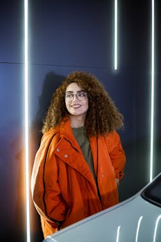 Stylish woman in orange jacket with curly hair and glasses in a modern indoor setting.