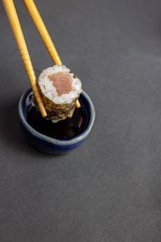 A detailed view of a tuna sushi roll being dipped into soy sauce with chopsticks.