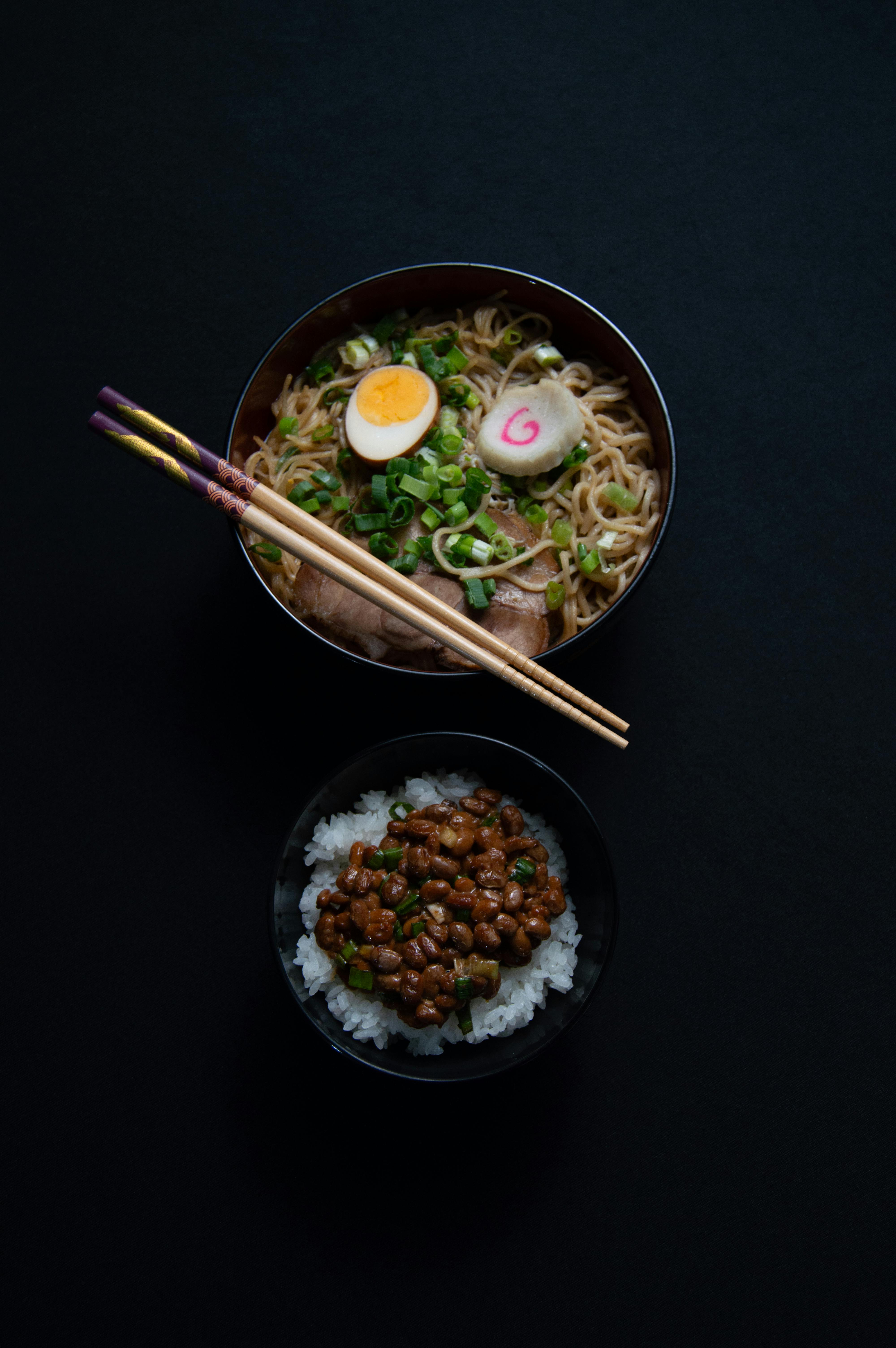 Flat lay of ramen and rice bowls with chopsticks on a dark surface, highlighting Japanese cuisine.