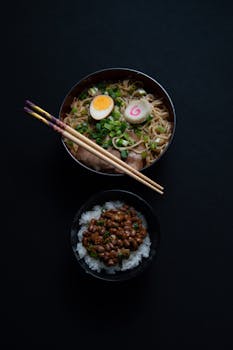 Flat lay of ramen and rice bowls with chopsticks on a dark surface, highlighting Japanese cuisine.