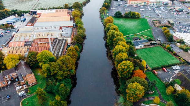 Aerial shot of a river bordered by trees and industrial buildings, showcasing autumn colors.