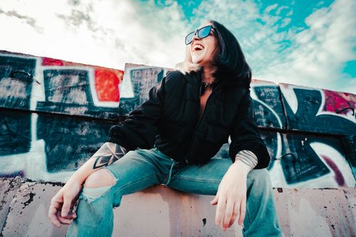 Free A woman in sunglasses laughs happily against a vibrant graffiti backdrop under a bright blue sky. Stock Photo
