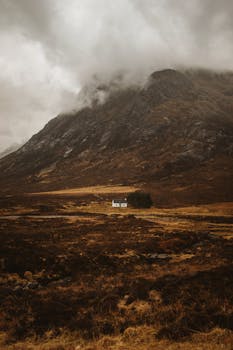 A solitary white house nestled in the dramatic landscape of Glencoe, Scotland, under a cloudy sky.