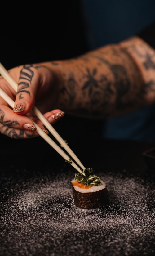 Free A tattooed hand uses chopsticks to pick up a sushi roll garnished with herbs on a dark background. Stock Photo