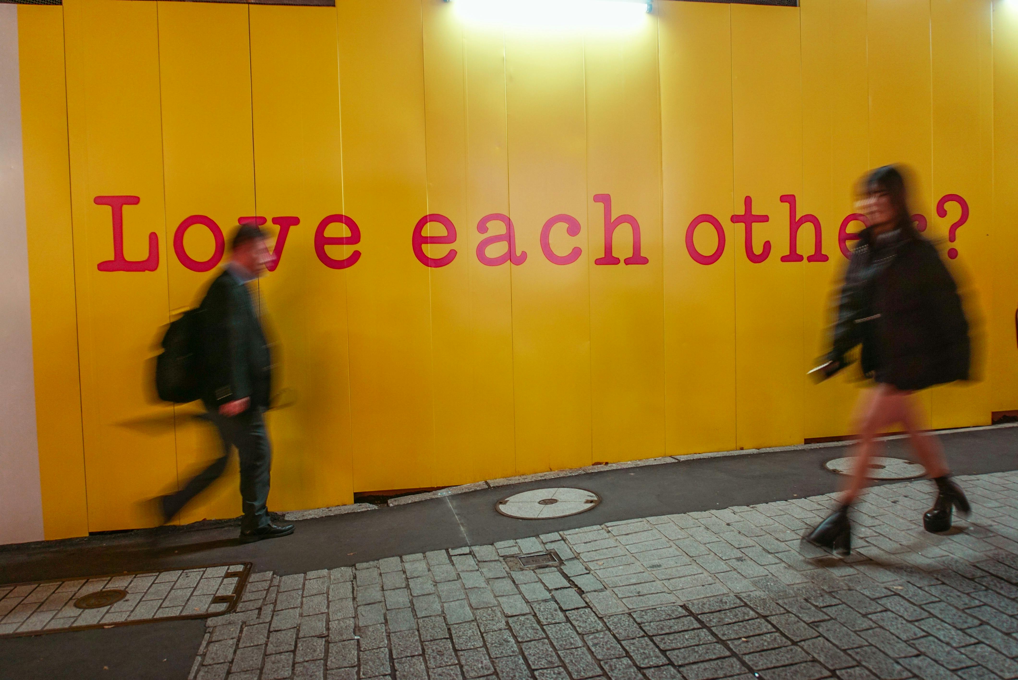 Blurred pedestrians pass by a bold yellow wall with 'Love each other?' in Shibuya, Tokyo.