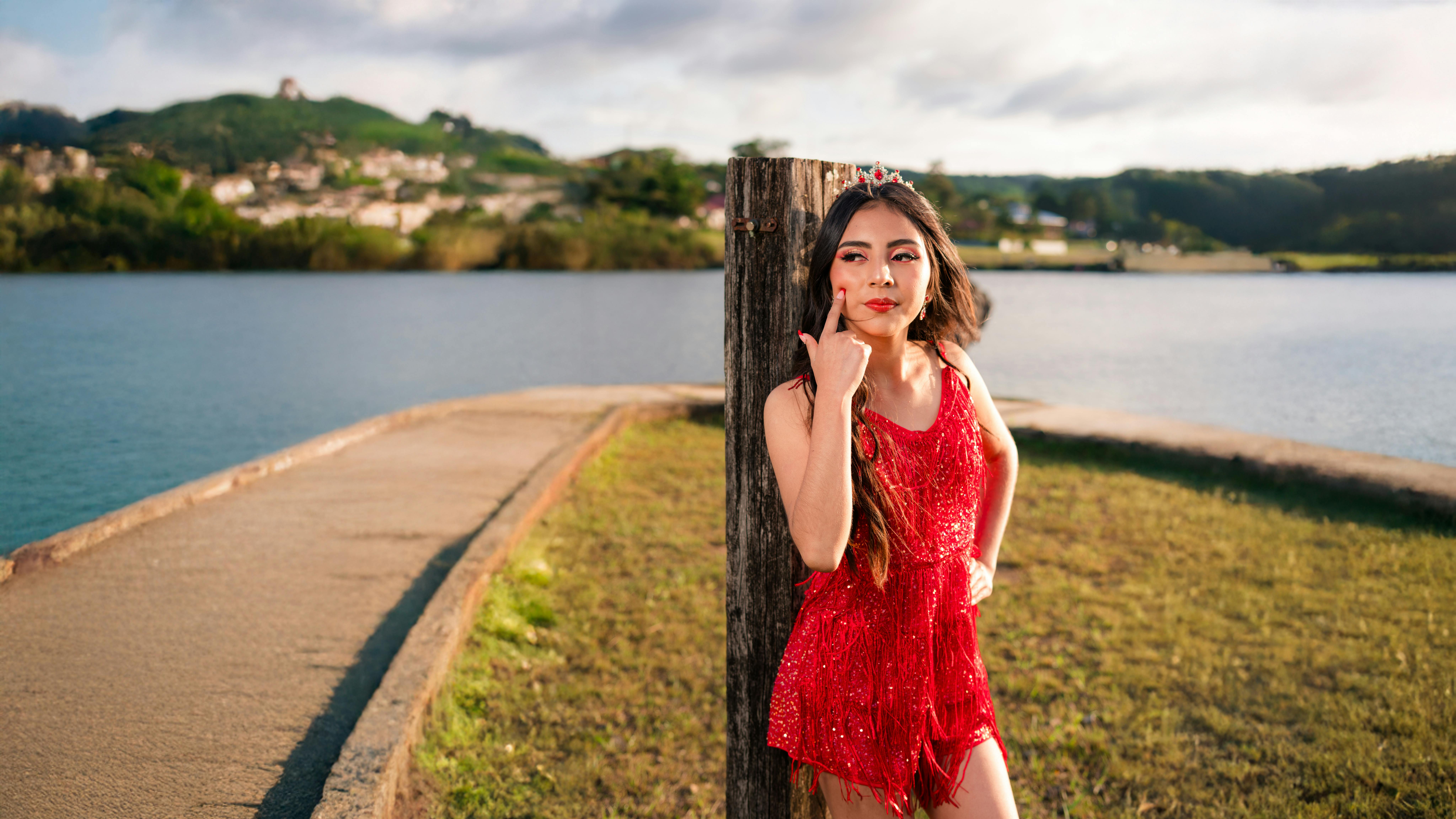 Young woman in a red dress stands by a lakeside, exuding confidence.