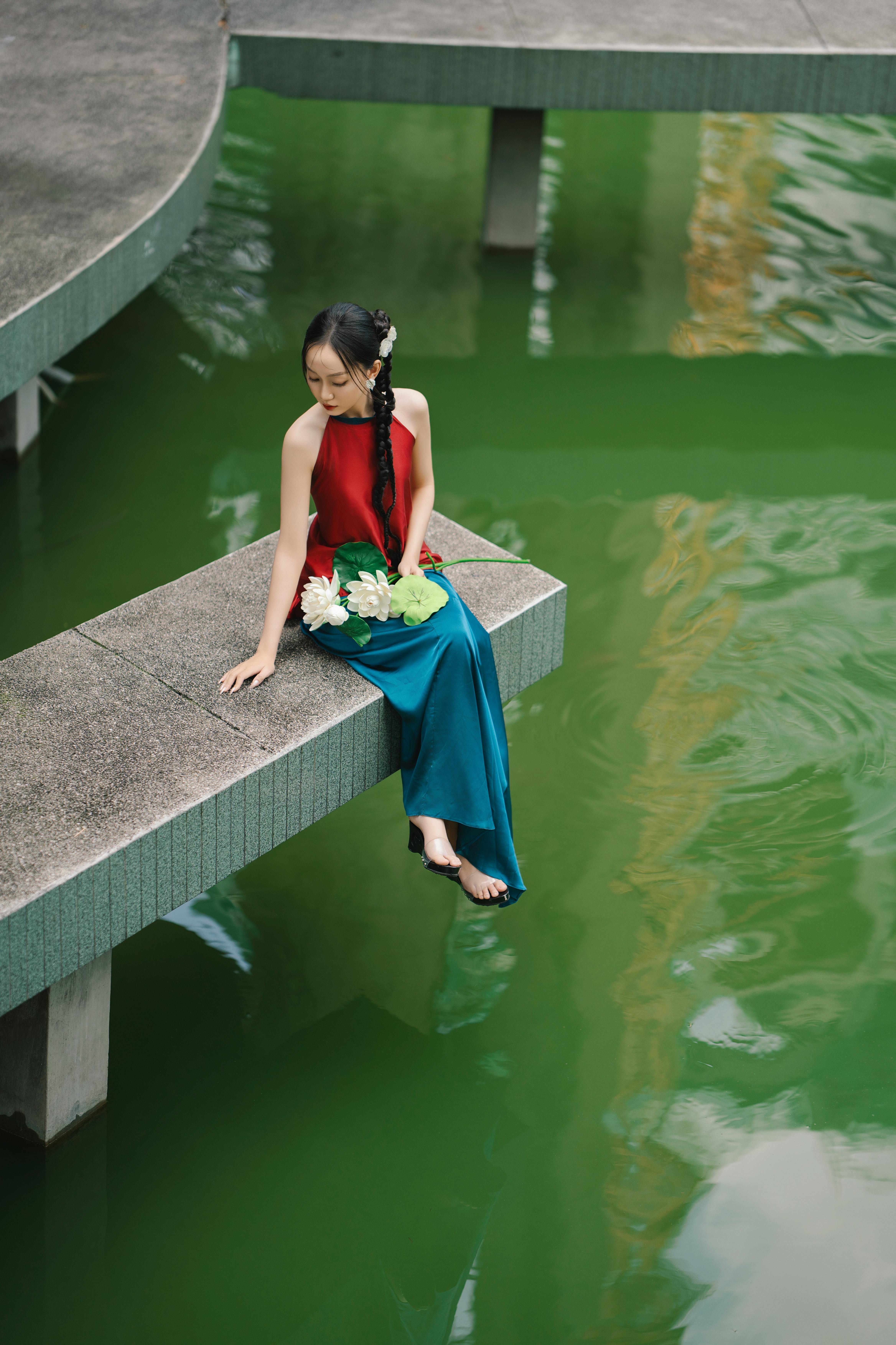 Elegant woman in a red dress sitting by a vibrant green pond, holding flowers. A serene outdoor scene.