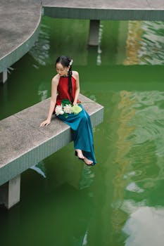 Elegant woman in a red dress sitting by a vibrant green pond, holding flowers. A serene outdoor scene.