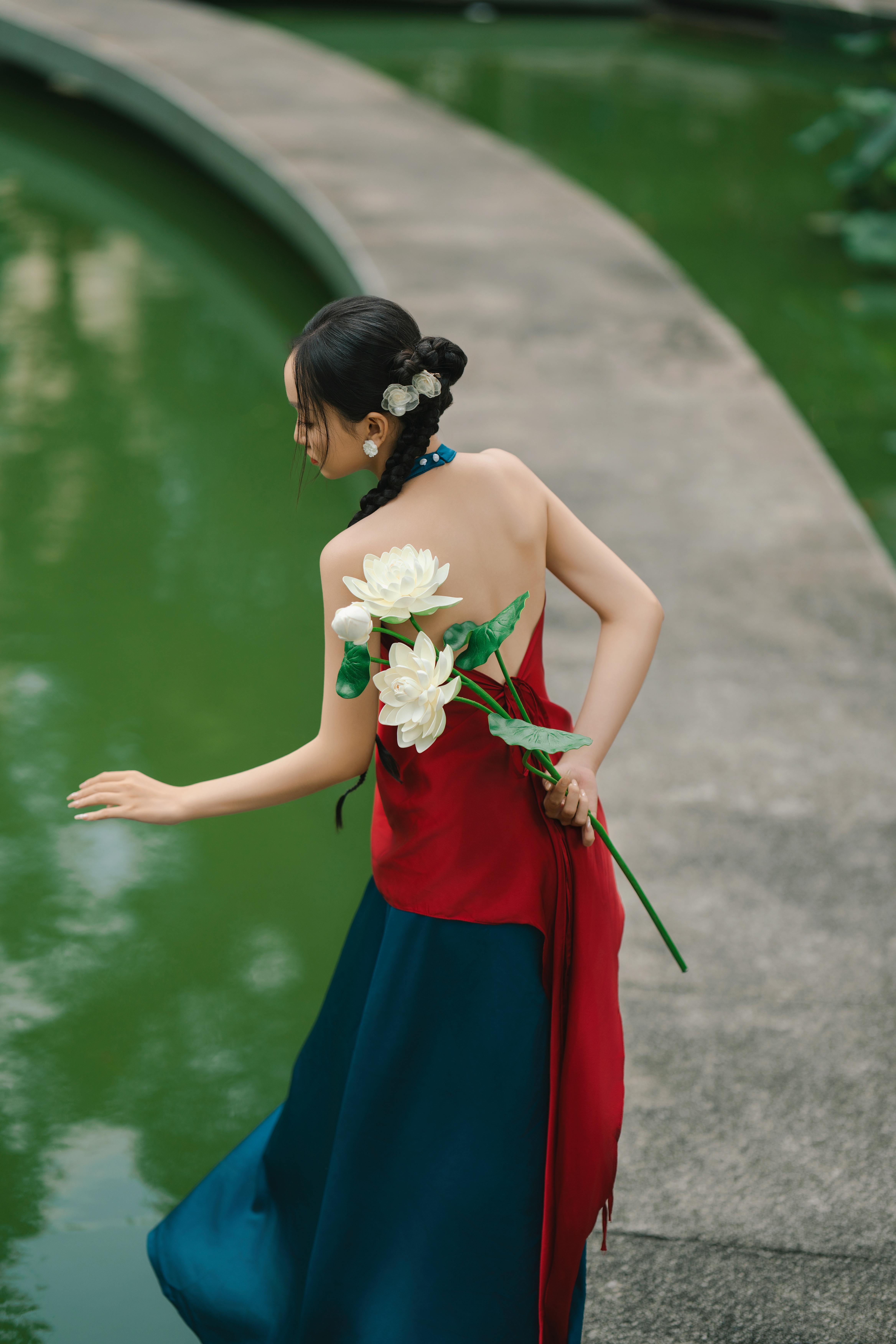 A woman in an elegant dress holding white flowers beside a serene green pond, exuding grace and calm.