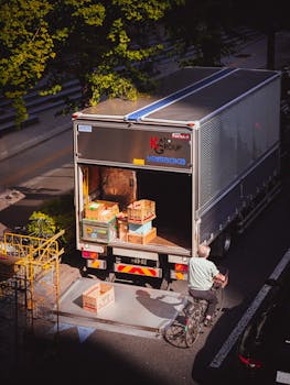 A delivery truck unloading boxes in a city street, with a bicyclist passing by on a sunny day.