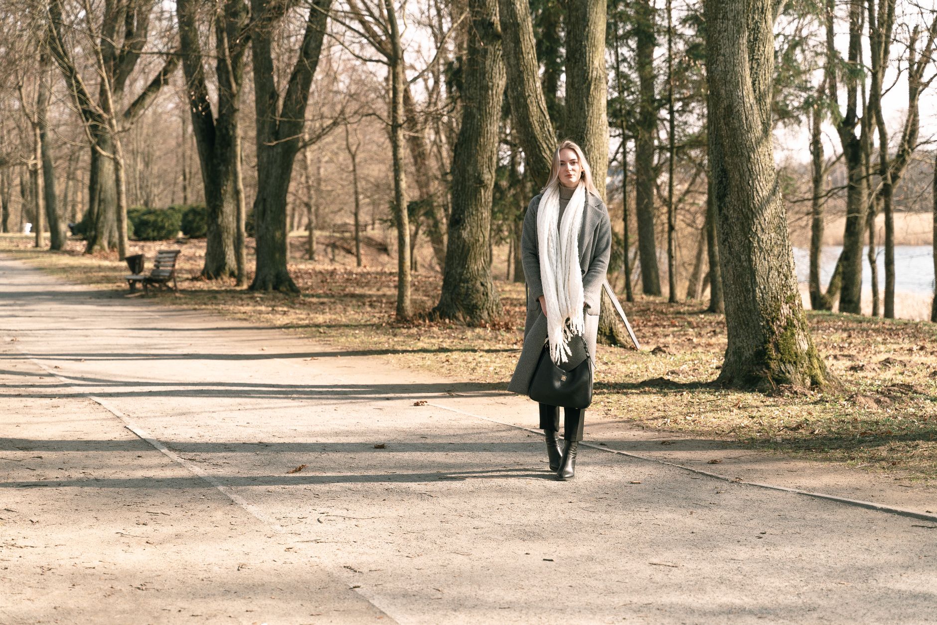 A woman in winter attire strolls through a serene tree-lined park on a sunny day.