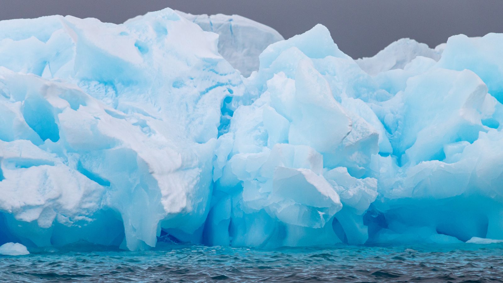 An immense blue-white iceberg rising from dark Antarctic waters beneath an overcast sky.