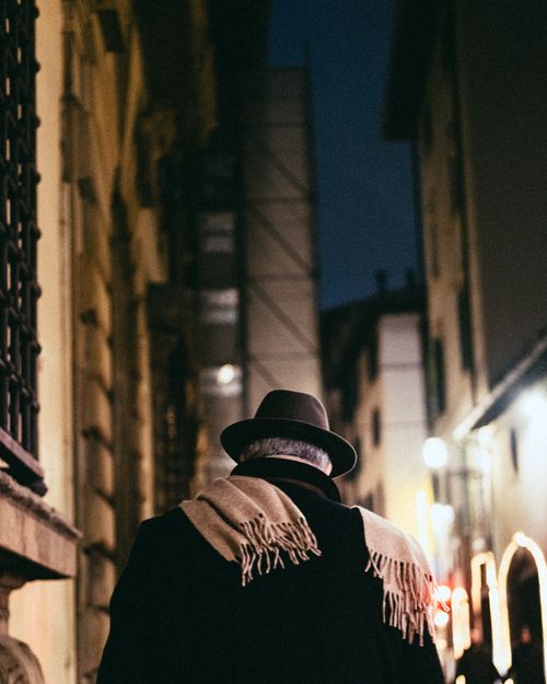 Free Back view of man in hat walking down lit urban street at night, creating intrigue. Stock Photo
