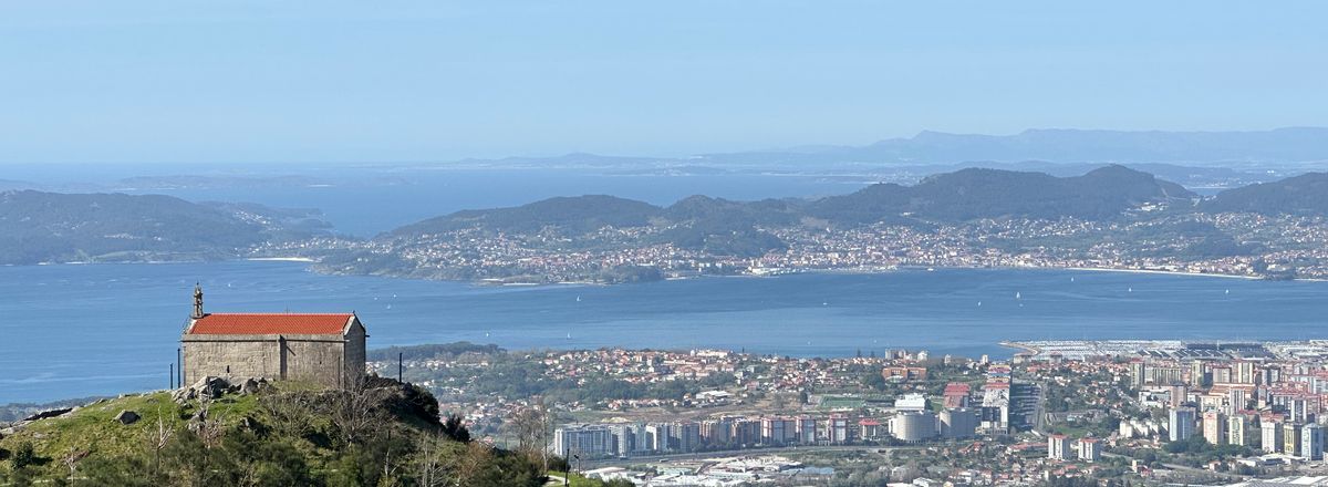 Aerial view of Vigo city, bay, and lush hills on a clear summer day, including a solitary building atop a hill.