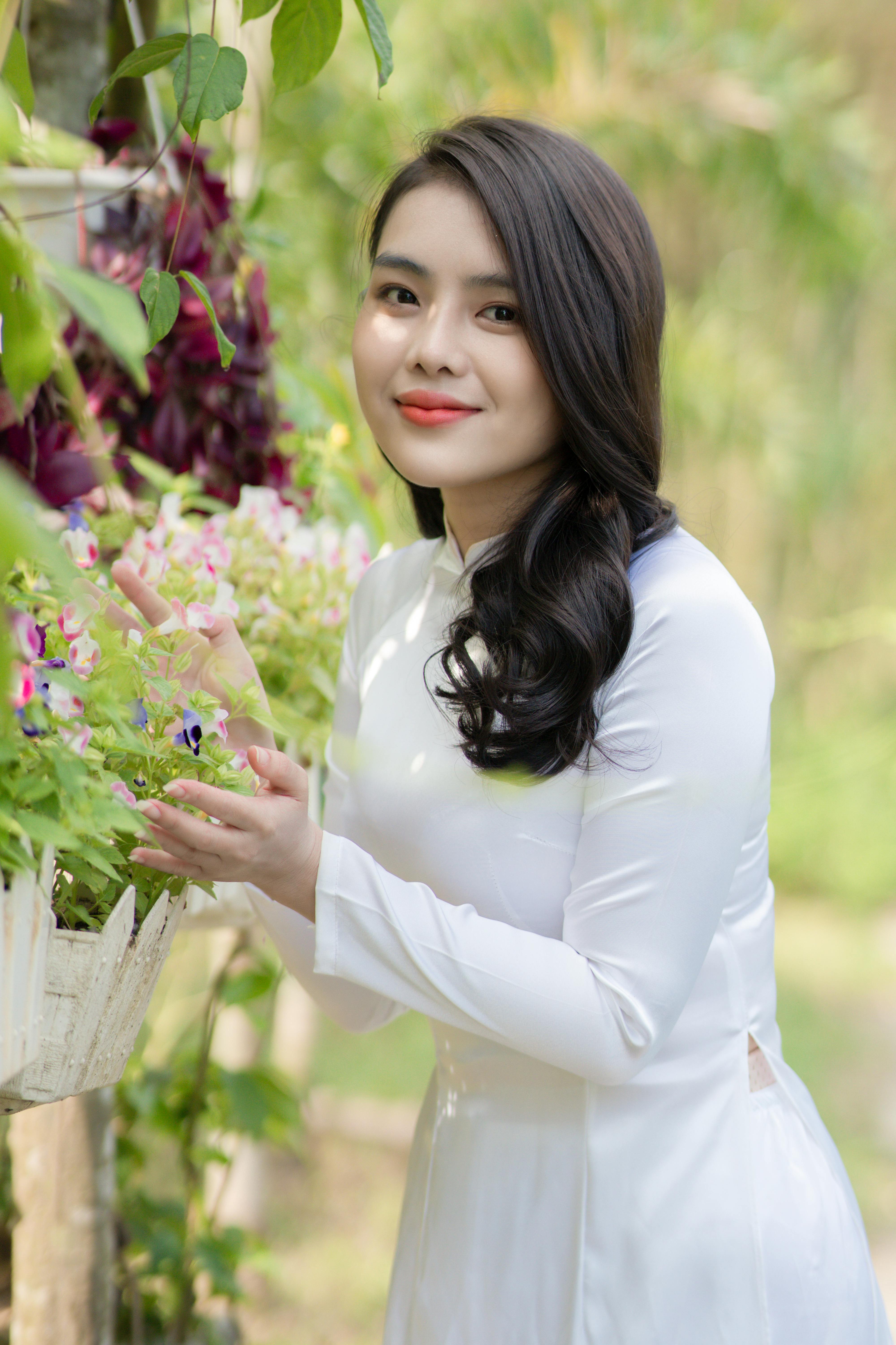 Asian woman in white Ao Dai smiling beside blossoming flowers outdoors.
