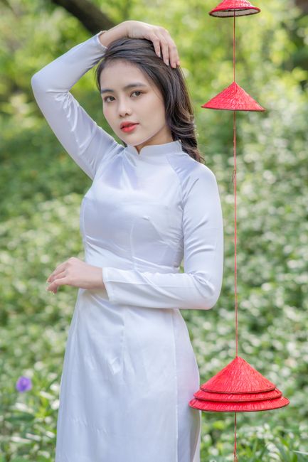 Elegant young Vietnamese woman in traditional white Ao Dai with red hats hanging, outdoors in nature