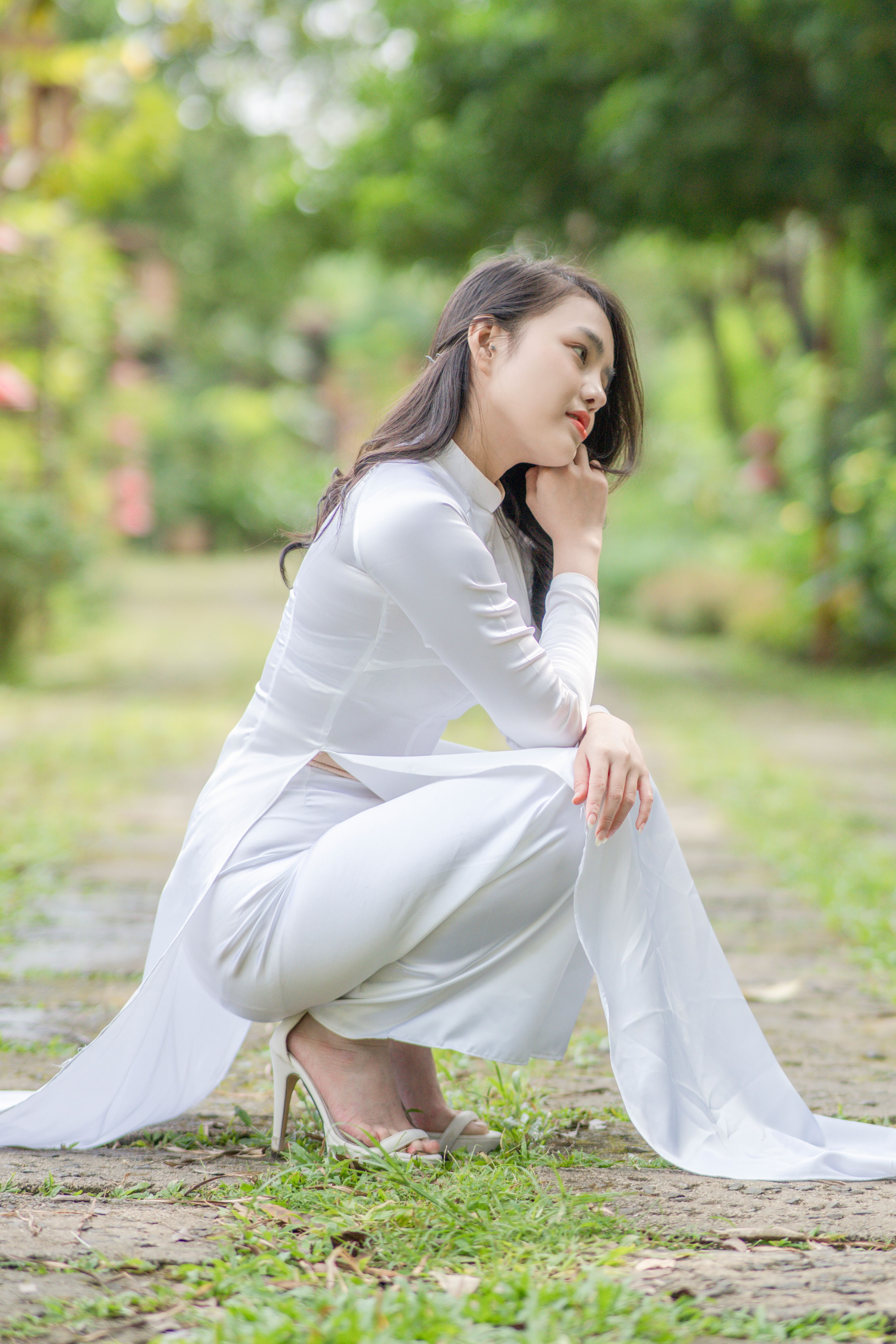 Elegant woman in white traditional dress crouching thoughtfully in a serene outdoor garden.