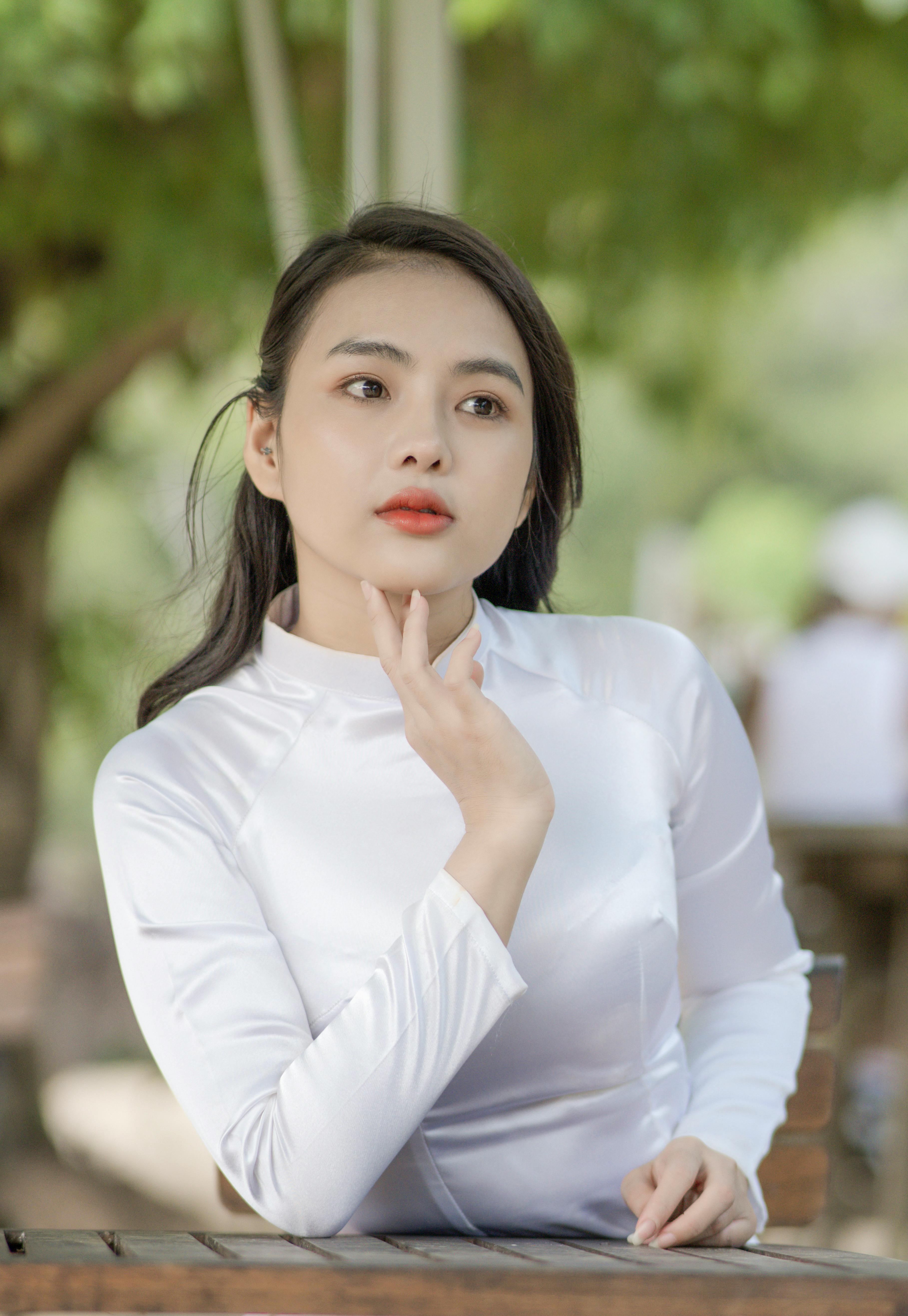 Ilmainen Tyylikäs nainen valkoisessa mekossa poseeraa mietteliäänä ulkona pehmeällä tarkennustaustalla. Kuvapankkikuva