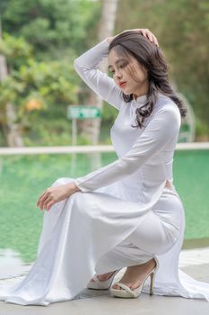 Woman in white traditional dress poses elegantly by a pool outdoors.