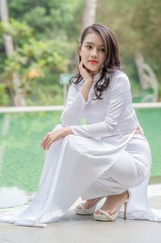 Woman in a white dress seated by a pool, exuding elegance and tranquility.