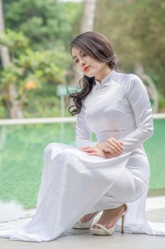 A woman wearing a traditional white Ao Dai dress poses gracefully by a pool, exuding elegance.