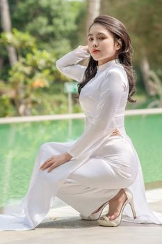 A woman dressed in a white Ao Dai poses elegantly by a serene green pool, capturing a graceful cultural moment.