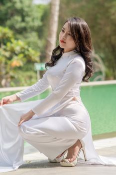 A graceful woman in a white Ao Dai kneels by a pool surrounded by greenery.