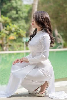 Woman in a flowing white traditional dress poses elegantly near a serene pool.