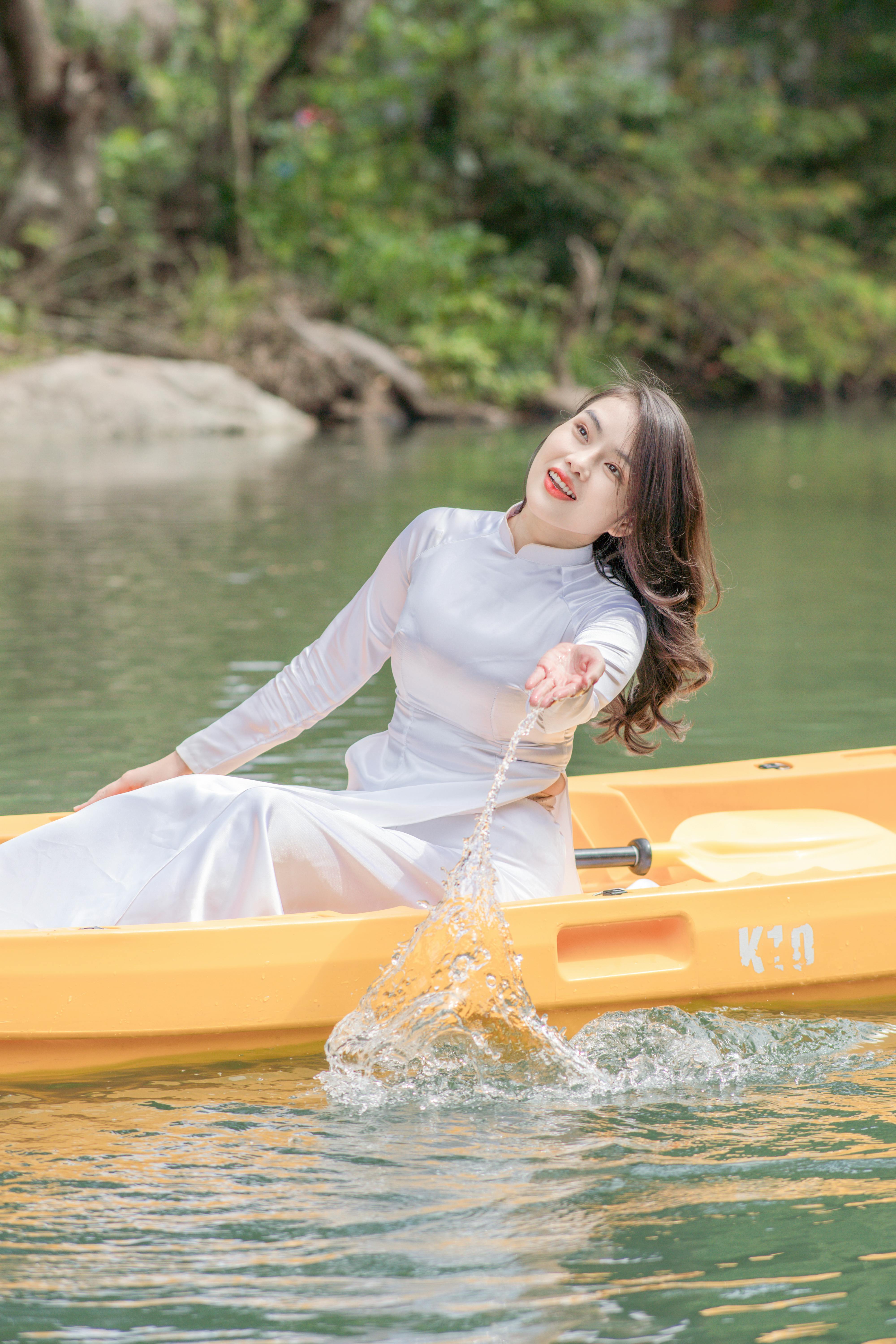 Woman in white traditional dress on a yellow kayak splashing water on a serene outdoor lake.