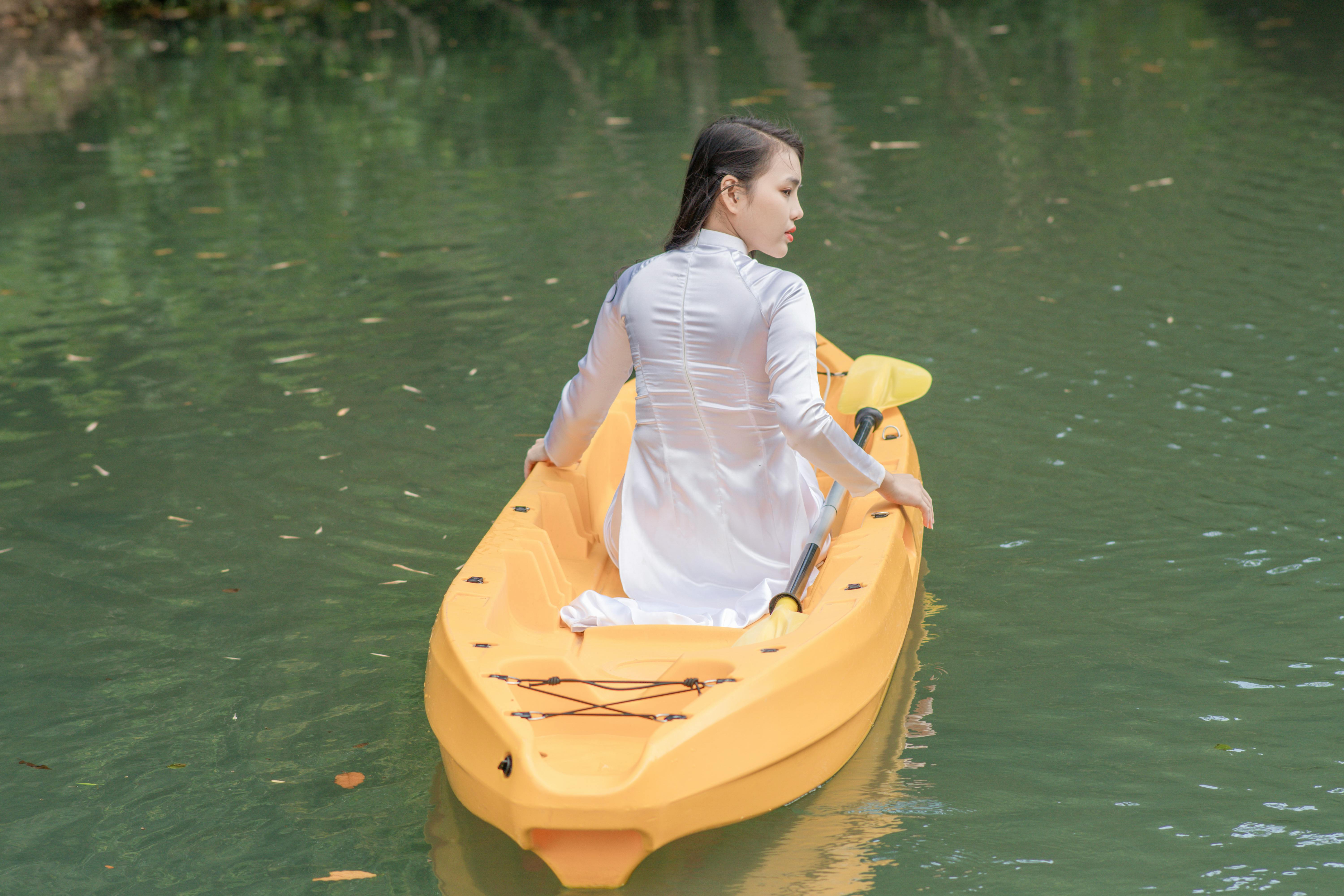 A woman in a white dress kayaking through calm green waters surrounded by nature.
